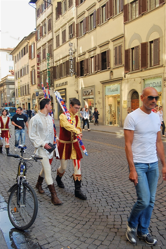 8 - A street scene in Florence, Italy, featuring participants in traditional historical Renaissance or medieval-style festive costumes carrying decorated staffs